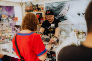 Verkaufsstand auf dem Elbenwald Festival, mit einem Verkäufer in einem schwarzen T-Shirt und einer Cap, der Merchandise wie Spielkarten und Figuren präsentiert. Besucher stöbern in den Angeboten, um einzigartige Sammlerstücke zu finden.