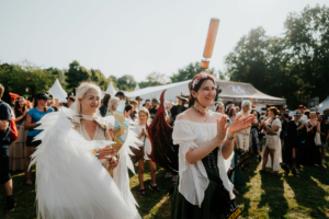 Zwei Teilnehmerinnen des Elbenwald Festivals in fantasievollen Kostümen, umgeben von einer jubelnden Menge. Die Frau links trägt beeindruckende Engelsflügel, während die Frau rechts begeistert applaudiert. Ein festlicher Moment voller Kreativität und Gemeinschaft.