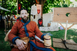 Die Roten Adler Ein Mann in einem orangefarbenen Gewand mit blauen Akzenten sitzt entspannt auf einem Stuhl, hält einen Holzkrug in der Hand. Im Hintergrund sind mittelalterliche Requisiten und Banner des Elbenwald Festivals zu sehen, das Musik und Fantasie verbindet.