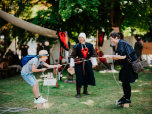 Die Roten Adler Zwei Teilnehmer des Elbenwald Festivals balancieren auf Holzblöcken und ziehen an einem Seil, während ein verkleideter Darsteller zuschaut. Die Szene zeigt die interaktive und kreative Atmosphäre des Festivals im Freien.