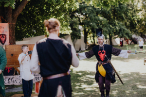 Die Roten Adler Ein Teilnehmer in historischer Kleidung führt eine Darbietung auf dem Elbenwald Festival vor, während ein Zuschauer mit einer Kamera filmt. Im Hintergrund sind Zelte und Festival-Elemente sichtbar, die die lebendige Atmosphäre des Events unterstreichen.
