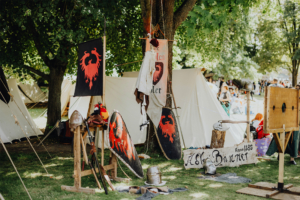 Die Roten Adler Bunt dekorierte Stände mit Schilden und Rüstungen auf dem Elbenwald Festival, umgeben von Bäumen und Zelten. Die Atmosphäre spiegelt den Mix aus Musik und Fantasy-Elementen wider, ideal für Fans von Rollenspielen und Cosplay.
