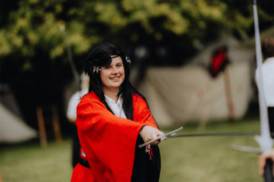 Schwertkampf mit Hammaborg Eine kostümierte Teilnehmerin in einem roten Kimono hält ein Schwert und lächelt fröhlich während des Elbenwald Festivals, umgeben von einer grünen Wiese und Zeltkulisse. Die Szene fängt die lebendige Atmosphäre des Festivals ein.