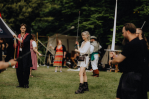 Schwertkampf mit Hammaborg Teilnehmer des Elbenwald Festivals in Kostümen üben mit Spielzeugschwertern auf einer Wiese. Im Hintergrund sind Zelte und andere Festivalbesucher zu sehen. Die Atmosphäre ist lebhaft und kreativ, ideal für Fans von Fantasy und Nerd-Kultur.
