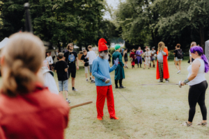 Schwertkampf Bunte Szene beim Elbenwald Festival: Teilnehmer in Kostümen interagieren auf einer Wiese, während einer im roten Hut und blauer Kleidung mit einem Schwert posiert. Im Hintergrund sind weitere Festivalbesucher und Stände zu sehen, die die lebendige Atmosphäre des Events unterstreichen.