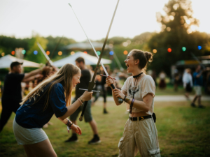 Schwertkampf Zwei Frauen kämpfen lachend mit Schwertattrappen auf dem Elbenwald Festival. Im Hintergrund sind weitere Festivalbesucher und bunte Lichter zu sehen, die die lebhafte Atmosphäre des Events unterstreichen.