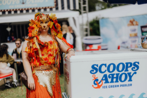 Eine festlich gekleidete Person mit einer bunten Maske aus Blättern steht neben einem Eisschrank von "Scoops Ahoy" auf dem Elbenwald Festival. Die lebhaften Farben und das kreative Kostüm spiegeln die einzigartige Atmosphäre des Festivals wider.