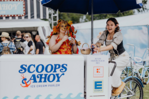 Zwei Frauen im Kostüm stehen an einem Eisstand beim Elbenwald Festival. Eine trägt ein auffälliges, blätterverziertes Outfit und genießt ein Eis, während die andere in einem historischen Kostüm ein Getränk serviert. Im Hintergrund sind Festivalbesucher zu sehen.