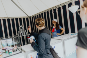 Scoops Ahoy Bunt dekorierter Stand auf dem Elbenwald Festival, mit einer lächelnden Verkäuferin in maritimer Kleidung. Im Hintergrund sind Stapel von Dosen und eine Auswahl an bunten Bechern zu sehen, während eine andere Person an der Theke steht.