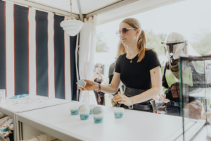 Scoops Ahoy Eine junge Frau in Sonnenbrille steht an einem Stand auf dem Elbenwald Festival und hält einen blauen Becher in der Hand. Auf dem Tisch sind weitere Becher und Merchandise-Artikel zu sehen. Im Hintergrund sind kostümierte Besucher des Festivals zu erkennen.