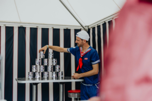Scoops Ahoy Ein Mann im Matrosenkostüm stapelt Dosen auf einem Tisch beim Elbenwald Festival. Der Hintergrund zeigt ein gestreiftes Zelt, das die kreative, nerdige Atmosphäre des Festivals unterstreicht. Spaß und Interaktion stehen im Vordergrund.