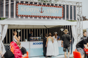 Scoops Ahoy Eisstand "Scoops Ahoy" auf dem Elbenwald Festival, mit Besuchern in kreativen Kostümen. Die festliche Atmosphäre wird durch das bunte Design und die fröhlichen Gesichter unterstrichen, während sie sich eine erfrischende Eiscreme gönnen.