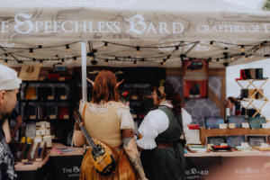 The Speechless Bard Zwei Besucherinnen mit fantasievollen Kostümen stehen am Stand "The Speechless Bard" auf dem Elbenwald Festival. Der Stand bietet handgefertigte Notizbücher und Accessoires für Rollenspieler und Fantasy-Fans, umgeben von bunten Lichtern und kreativen Produkten.
