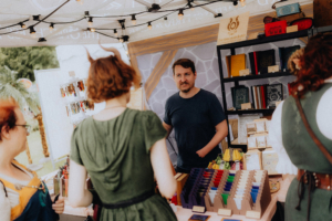 The Speechless Bard Ein Stand auf dem Elbenwald Festival mit einem Verkäufer, der handgefertigte Notizbücher und kreative Accessoires präsentiert. Besucherinnen stöbern in den bunten Angeboten und genießen die entspannte Atmosphäre des Festivals.