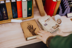 The Speechless Bard Ein handgefertigtes Notizbuch mit einem kunstvollen Handdesign liegt auf einem Tisch, umgeben von bunten, dekorativen Büchern und Informationsflyern. Die Atmosphäre spiegelt die kreative und fantasievolle Stimmung des Elbenwald Festivals wider.