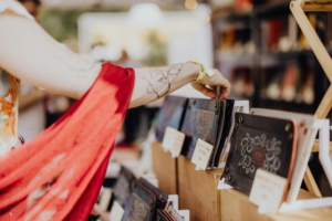 The Speechless Bard Eine Person mit einem roten Umhang greift nach einem Notizbuch auf einem Marktstand beim Elbenwald Festival, umgeben von handgefertigten Kunstwerken und Merchandise, die das kreative und nerdige Ambiente der Veranstaltung widerspiegeln.