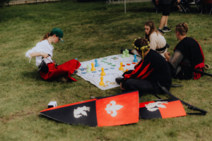 Gruppe von Teilnehmern beim Elbenwald Festival, die auf einer Wiese ein Brettspiel spielen. Im Vordergrund sind bunte Spielfiguren und Schilde zu sehen, während die Festivalbesucher in fantasievollen Kostümen konzentriert am Spiel teilnehmen.