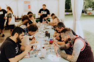 Teilnehmer des Elbenwald Festivals sitzen an einem Tisch und bemalen Miniaturen. Die kreative Atmosphäre fördert den Austausch unter Nerds und Fans, während verschiedene Farben und Materialien auf dem Tisch verteilt sind.