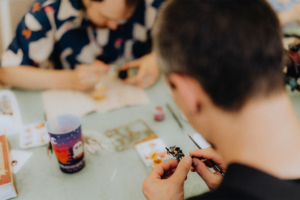 Ein Teilnehmer des Elbenwald Festivals malt ein Miniaturmodell, während im Hintergrund ein weiterer Bastler beschäftigt ist. Auf dem Tisch sind Malutensilien und ein Becher mit fantasievollem Design sichtbar, die die kreative Atmosphäre des Festivals unterstreichen.