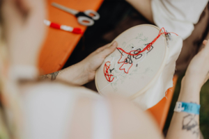 Stitchcraft Eine Person hält einen Stickrahmen mit einem farbenfrohen Stickmuster in der Hand. Im Hintergrund sind Scheren und Garn sichtbar. Die Szene vermittelt die kreative Atmosphäre des Elbenwald Festivals, einem beliebten Event für Nerd-Kultur und Kunst.