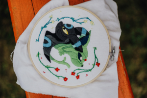 Stitchcraft Detailaufnahme eines handgestickten Kunstwerks, das eine fantasievolle Kreatur in lebendigen Farben zeigt, umgeben von floralen Mustern. Die Stickerei ist auf einem Stickrahmen gespannt und wird auf einem Holzuntergrund präsentiert, ideal für kreative Nerd-Kunst.