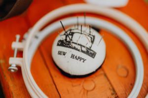 Stitchcraft Nähzubehör auf einem Holztisch beim Elbenwald Festival, mit einem Nadelkissen, das die Aufschrift "SEW HAPPY" trägt, umgeben von Nähnadeln und einem Stickrahmen. Ideal für kreative Bastelarbeiten und Handarbeiten auf dem Festival.