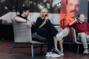 Stranger Things Synchro-Talk Podiumsdiskussion auf dem Elbenwald Festival: Drei Sprecher sitzen auf gemütlichen Stühlen, während einer von ihnen ein Mikrofon hält. Im Hintergrund ist ein Plakat mit einem Porträt zu sehen. Die Atmosphäre ist entspannt und kreativ.