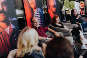 Stranger Things Synchro-Talk mit Peter Flechtner, Franciska Friede und Martin Sabel Podiumsdiskussion auf dem Elbenwald Festival mit mehreren Sprechern, die in gemütlichen Stühlen sitzen. Im Hintergrund sind große Leinwände mit Porträts von Charakteren zu sehen. Die Atmosphäre ist entspannt und kreativ, ideal für Fans von Musik und Nerdkultur.
