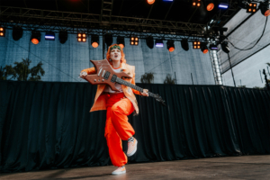 Stranger Things Cosplay Contest Eine energiegeladene Musikerin in orangefarbener Kleidung spielt eine auffällige Gitarre auf der Bühne des Elbenwald Festivals, umgeben von bunten Lichtern und einer begeisterten Menge. Ihre Performance vereint Musik und Nerd-Kultur in einem einzigartigen Erlebnis.
