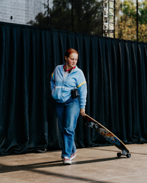 Stranger Things Cosplay Contest Junge Frau mit rotem Haar und Gesichtsbemalung, trägt eine blaue Jacke und Jeans, während sie ein Skateboard hält. Sie bewegt sich auf einer Festivalbühne beim Elbenwald Festival, umgeben von schwarzen Vorhängen.