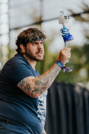 Stranger Things Cosplay Contest Ein tätowierter Mann auf der Bühne des Elbenwald Festivals hält einen kreativen, selbstgemachten Stock mit verschiedenen bunten Elementen. Die Atmosphäre ist lebhaft und zeigt die Verbindung von Musik und Nerd-Kultur auf diesem einzigartigen Event.