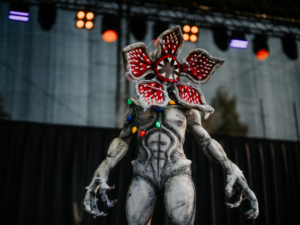 Stranger Things Cosplay Contest Kreatives Kostüm eines Monsters mit einer blühenden Kopfstruktur und bunten Lichtern, präsentiert auf der Bühne des Elbenwald Festivals. Die Atmosphäre ist lebhaft und fantasievoll, mit einem Fokus auf Nerd- und Popkultur-Elemente.