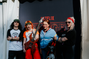 Vier Cosplayer posieren vor einem Netflix-Stand auf dem Elbenwald Festival. Sie tragen stylische Outfits, darunter ein T-Shirt des Hellfire Clubs und halten eine E-Gitarre sowie ein Skateboard. Die Atmosphäre ist lebhaft und kreativ.
