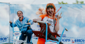 Stranger Things Cosplay Zwei Festivalbesucher beim Elbenwald Festival, einer mit einer stylischen E-Gitarre in Form eines Sterns und der andere auf einem kleinen Fahrrad. Bunte Kleidung und eine fröhliche Atmosphäre spiegeln die nerdige Stimmung des Events wider.