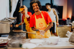 Surfer Boy Pizza Ein junger Mann mit lockigen Haaren und einem freundlichen Lächeln trägt ein gelbes Schürze und bereitet Pizza in einem Food-Stand beim Elbenwald Festival vor. Im Hintergrund sind weitere Mitarbeiter zu sehen, die ebenfalls an der Zubereitung von Speisen arbeiten.