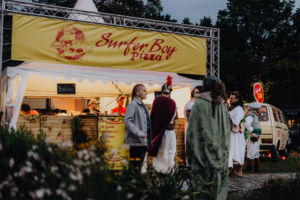 Surfer Boy Pizza Bunte Szenerie am Elbenwald Festival mit dem Verkaufsstand "Surfer Boy Pizza". Besucher in kreativen Kostümen stehen Schlange, während das Menü mit verschiedenen Pizzavarianten präsentiert wird. Gemütliche Atmosphäre und vielfältige kulinarische Angebote.