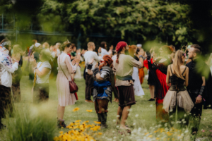 Historische Tänze Bunt gekleidete Festivalbesucher interagieren auf dem Elbenwald Festival, umgeben von Blumen und einer lebendigen Atmosphäre. Die Szene zeigt Kostüme und kreative Outfits, die die Vielfalt der Nerd- und Fantasiekultur feiern.
