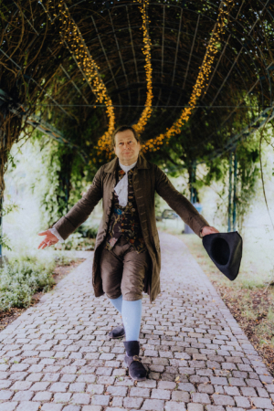 Ein Mann in historischer Kleidung mit einem Zylinderhut läuft durch einen mit Lichtern geschmückten Gartenweg. Die Szenerie strahlt eine festliche Atmosphäre aus, passend zum Elbenwald Festival, das Musik und nerdige Kultur vereint.