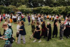 Historische Tänze Bunte Menschenmenge beim Elbenwald Festival, verkleidet in fantasievollen Kostümen, tanzt und feiert auf einer Wiese. Die Atmosphäre ist lebhaft, mit einer Mischung aus Musik und kreativen Darstellungen, die den nerdigen Charakter des Festivals unterstreichen.