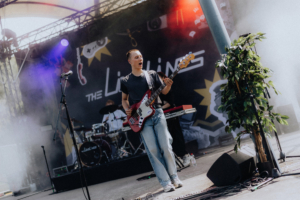 The Livelines Ein Musiker spielt Bass auf der Bühne beim Elbenwald Festival. Im Hintergrund sind Schlagzeug und Keyboard sichtbar, während bunte Lichter und Nebel die Atmosphäre des Events betonen. Die Band "The Linings" sorgt für eine energiegeladene Stimmung.