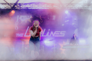 The Livelines Die Sängerin der Band The Lainlines tritt auf der Bühne des Elbenwald Festivals auf, umgeben von Nebel und bunten Lichtern. Die energiegeladene Atmosphäre vereint Musik und nerdige Kultur, während die Zuschauer begeistert mitfeiern.