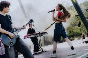 The Livelines Auf der Bühne des Elbenwald Festivals performt eine Sängerin mit Mikrofon, während ein Gitarrist im Vordergrund spielt. Eine Keyboarderin ergänzt die Band. Die Atmosphäre ist lebhaft, umgeben von Nebel und einer begeisterten Menge.