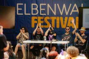 The Nerds Fünf Teilnehmer applaudieren auf der Bühne des Elbenwald Festivals, umgeben von einer bunten Menge. Der Hintergrund zeigt das Festival-Logo, während die Atmosphäre lebhaft und festlich ist. Ein Moment der Begeisterung und Gemeinschaft auf dem Event.