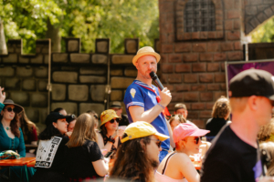 Olaf Unzel Ein Moderator in einem maritimen Kostüm spricht mit einem Mikrofon zu einer bunten Menge von Festivalbesuchern, die in einem sonnigen, festlichen Ambiente am Elbenwald Festival sitzen. Im Hintergrund sind Dekorationen und eine Mauer aus Stein sichtbar.