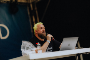Tobias M. Eckrich Ein Moderator mit gelben Haaren spricht am Mikrofon auf der Bühne des Elbenwald Festivals, während ein Laptop und eine Flasche Wasser auf dem Tisch stehen. Die Veranstaltung kombiniert Musik und Nerd-Kultur und zieht zahlreiche Fans an.