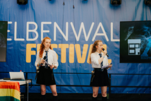 Typisch Ravenclaw Zwei Moderatorinnen in Schuluniform stehen auf der Bühne des Elbenwald Festivals und präsentieren vor einem blauen Banner. Im Hintergrund ist ein Bildschirm mit einem grafischen Element zu sehen. Die Atmosphäre ist kreativ und lebhaft, ideal für Nerd-Kultur.