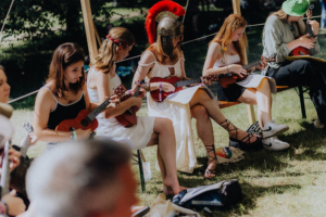 Ukulele Workshop Eine Gruppe junger Frauen spielt Ukulele in festlicher Kleidung, darunter eine mit einem römischen Helm. Die Szene findet im Freien während des Elbenwald Festivals statt, wo Musik und kreative Ausdrucksformen im Mittelpunkt stehen.