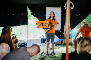 Eine Musikerin mit Ukulele steht auf einer Bühne im Zelt des Elbenwald Festivals und singt, während das Publikum ihr aufmerksam zuhört. Im Hintergrund sind Notenblätter und Instrumente sichtbar, die die kreative Atmosphäre des Festivals unterstreichen.