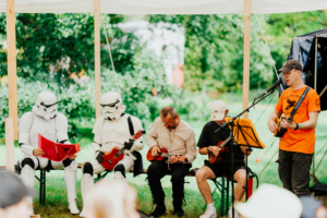 Ukulele Workshop: Musiker und Stormtrooper auf dem Elbenwald Festival: Zwei Stormtrooper sitzen mit Ukulelen auf einer Bühne, während ein Sänger mit Gitarre auftritt. Die entspannte Atmosphäre und die kreative Mischung aus Musik und Nerdkultur ziehen das Publikum an. Foto: Robin Schmiedebach