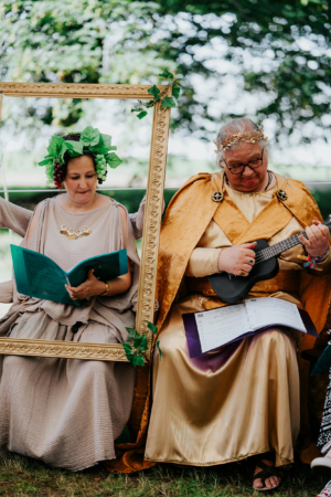 Zwei Frauen in fantasievollen Kostümen sitzen beim Elbenwald Festival, eine mit Traubenkranz und Buch, die andere mit einer Ukulele und Noten. Umrahmt von einem goldenen Bilderrahmen, verkörpern sie kreative Charaktere in einer lebhaften Festivalatmosphäre.